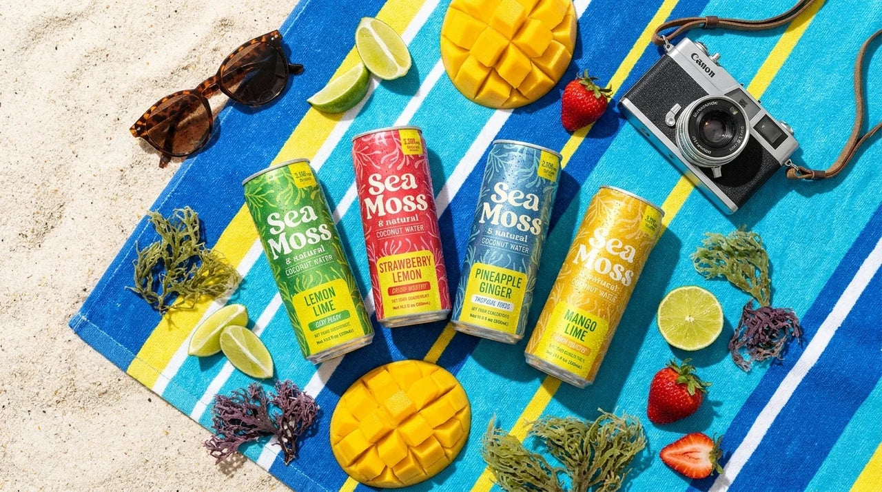 All four Da Bomb Sea Moss Coconut Water flavors on a beach towel with fresh fruit and wildcrafted sea moss